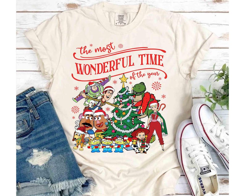 May include: Cream-colored t-shirt featuring a festive design with the text "The Most Wonderful Time of the Year" in red. The graphic includes cartoon characters, a Christmas tree, and snowflakes, creating a holiday theme.