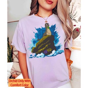 May include: A light purple t-shirt featuring a cartoon illustration of a sea creature on a rock with blue water and a splash effect. The text "Orchid Only With Comfort Color" is in a yellow box at the bottom.