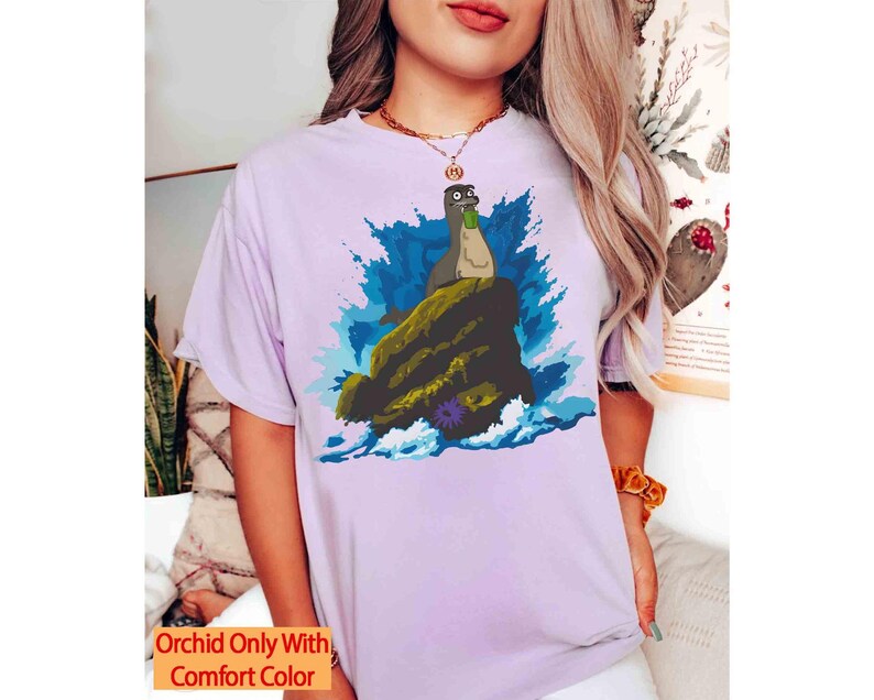 May include: A light purple t-shirt featuring a cartoon illustration of a sea creature on a rock with blue water and a splash effect. The text "Orchid Only With Comfort Color" is in a yellow box at the bottom.