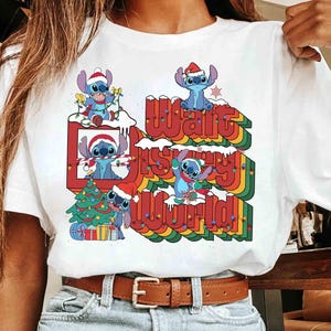 K&ouml;nnte beinhalten: Wei&szlig;es T-Shirt mit einem farbenfrohen Aufdruck der Figur Stitch mit Weihnachtsm&uuml;tze und dem Text "Walt Disney World" in einem Retro-Regenbogen-Design. Das Design beinhaltet einen Weihnachtsbaum und Schnee.