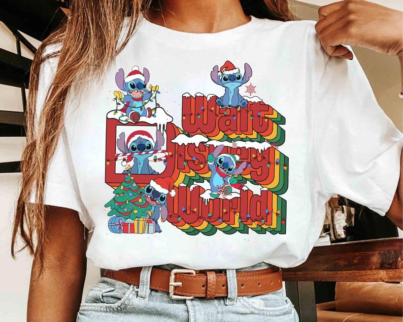 K&ouml;nnte beinhalten: Wei&szlig;es T-Shirt mit einem farbenfrohen Aufdruck der Figur Stitch mit Weihnachtsm&uuml;tze und dem Text "Walt Disney World" in einem Retro-Regenbogen-Design. Das Design beinhaltet einen Weihnachtsbaum und Schnee.