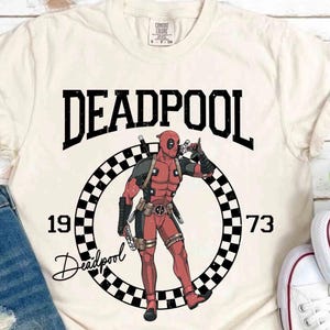 Puede incluir: Camiseta color crema con un gr&aacute;fico de Deadpool. El dise&ntilde;o incluye la palabra "DEADPOOL" sobre un c&iacute;rculo a cuadros con los n&uacute;meros "1973" y la ilustraci&oacute;n del personaje. La camiseta se combina con vaqueros azules y zapatillas blancas.