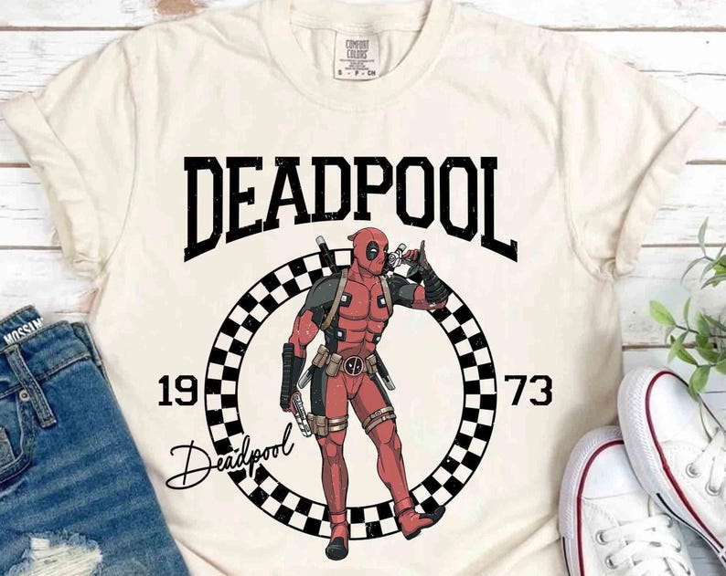 Puede incluir: Camiseta color crema con un gr&aacute;fico de Deadpool. El dise&ntilde;o incluye la palabra "DEADPOOL" sobre un c&iacute;rculo a cuadros con los n&uacute;meros "1973" y la ilustraci&oacute;n del personaje. La camiseta se combina con vaqueros azules y zapatillas blancas.