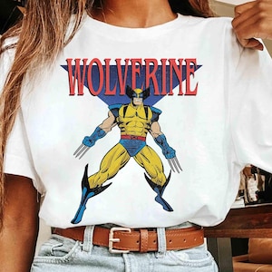 May include: A white short-sleeved t-shirt featuring a graphic print of the comic book character Wolverine in a yellow and blue costume with metal claws. The word WOLVERINE is printed in large red letters above the character, set against a blue triangle.