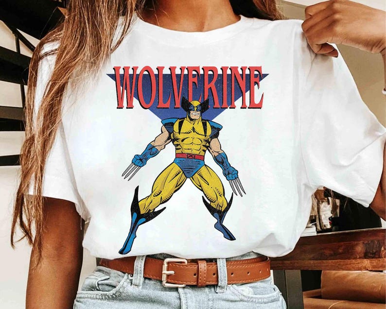 May include: A white short-sleeved t-shirt featuring a graphic print of the comic book character Wolverine in a yellow and blue costume with metal claws. The word WOLVERINE is printed in large red letters above the character, set against a blue triangle.