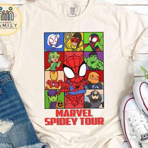 May include: Cream-colored t-shirt featuring a colorful grid of Marvel characters, including Spider-Man. The shirt has the text "MARVEL SPIDEY TOUR" in red. The shirt is paired with blue denim shorts and white sneakers.