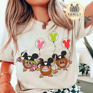 May include: A cream-colored t-shirt featuring a cartoon design of three characters with Mickey Mouse ears, holding weapons and balloons. The balloons are pink, green, and red. The shirt has the text "HAPPY FAMILY" in a circle.