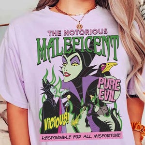 May include: A lilac t-shirt with a graphic of Maleficent from Disney's Sleeping Beauty. The design includes the text "THE NOTORIOUS MALEFICENT" and "PURE EVIL", along with other characters and text. The shirt is a crew neck.