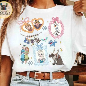 Op de afbeelding: Een wit t-shirt met korte mouwen en een kleurrijke grafische print van personages uit de film Frozen. Het ontwerp bevat Anna, Elsa, Kristoff, Olaf en Sven, samen met decoratieve kaders, sneeuwvlokken en de tekst Frozen op een banner.