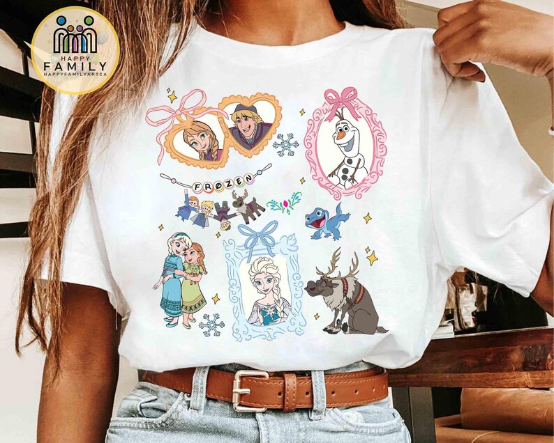 Op de afbeelding: Een wit t-shirt met korte mouwen en een kleurrijke grafische print van personages uit de film Frozen. Het ontwerp bevat Anna, Elsa, Kristoff, Olaf en Sven, samen met decoratieve kaders, sneeuwvlokken en de tekst Frozen op een banner.