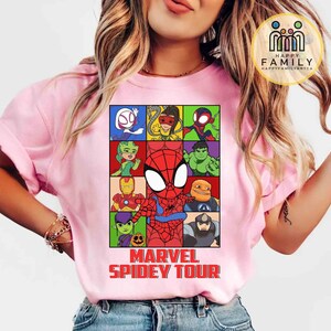 May include: Pink t-shirt featuring a colorful graphic of Marvel characters, including Spider-Man, Iron Man, and Hulk. The design is set in a grid pattern with the text "MARVEL SPIDEY TOUR" in red at the bottom. The shirt has short sleeves.