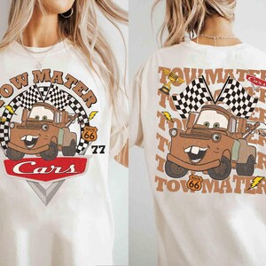 May include: Cream-colored t-shirt featuring a cartoon image of Tow Mater from the movie Cars. The design includes the text "Tow Mater," a checkered flag, and the Route 66 logo. The front and back of the shirt are visible.