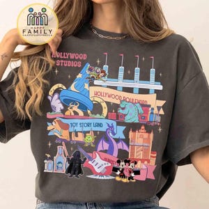 Può includere: T-shirt grigio scuro con una grafica colorata delle attrazioni e dei personaggi di Hollywood Studios. Il design include il testo "Hollywood Studios" e "Toy Story Land". La maglietta ha una vestibilità ampia.