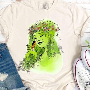 May include: Cream-colored t-shirt featuring a green illustration of a woman with a floral crown and a small figure in her hand. The design has a watercolor style. The shirt is paired with denim shorts and white sneakers.