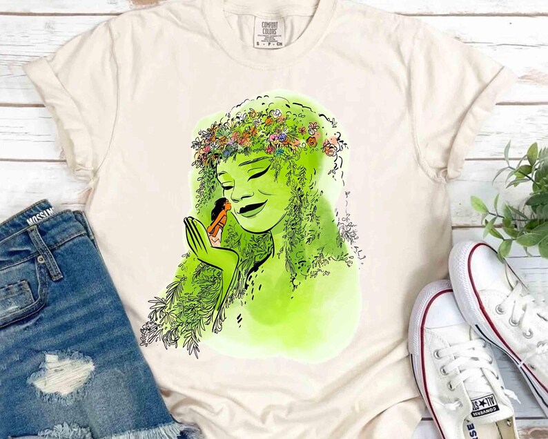 May include: Cream-colored t-shirt featuring a green illustration of a woman with a floral crown and a small figure in her hand. The design has a watercolor style. The shirt is paired with denim shorts and white sneakers.