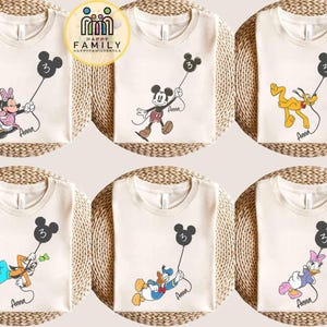 Puede incluir: Camisetas color crema con varios personajes de Disney sosteniendo globos negros con forma de Mickey Mouse con el n&uacute;mero 3. Cada camiseta tiene el nombre de un personaje escrito en cursiva. Los personajes incluyen Minnie, Mickey, Pluto, Goofy, Donald y Daisy.