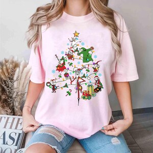 以下が含まれることがあります： ナイトメアー・ビフォア・クリスマスのキャラクターが描かれたクリスマスツリーのデザインの、明るいピンク色のTシャツ。ツリーはカラフルなオーナメントとライトで飾られています。クルーネックの半袖Tシャツです。