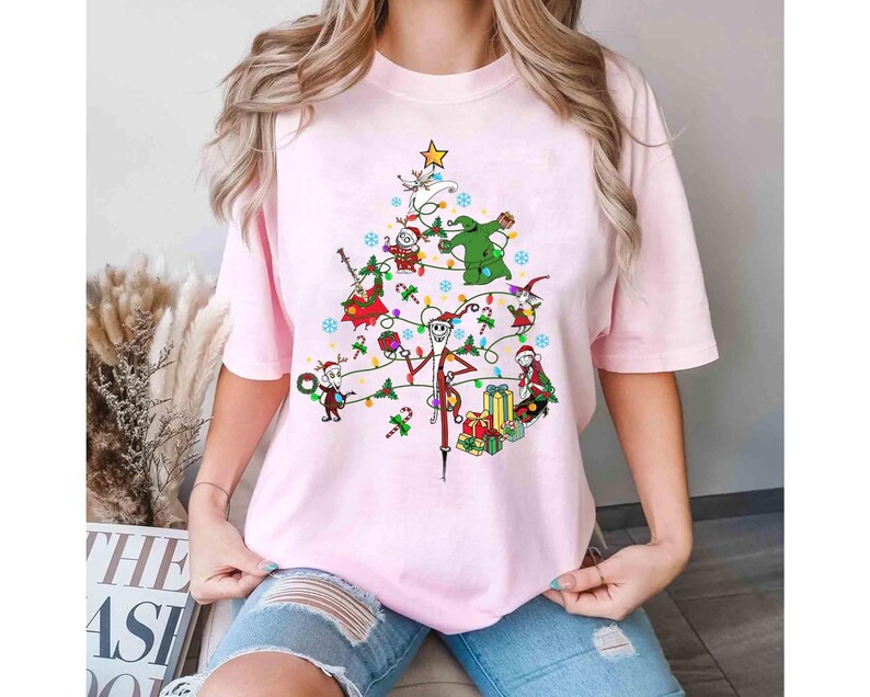 以下が含まれることがあります： ナイトメアー・ビフォア・クリスマスのキャラクターが描かれたクリスマスツリーのデザインの、明るいピンク色のTシャツ。ツリーはカラフルなオーナメントとライトで飾られています。クルーネックの半袖Tシャツです。
