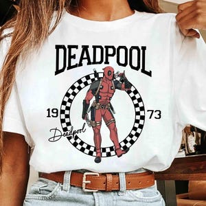 Puede incluir: Camiseta blanca con un dise&ntilde;o de Deadpool. El dise&ntilde;o incluye la palabra "DEADPOOL" sobre un c&iacute;rculo a cuadros con la ilustraci&oacute;n del personaje, los n&uacute;meros "1973" y la firma de Deadpool.