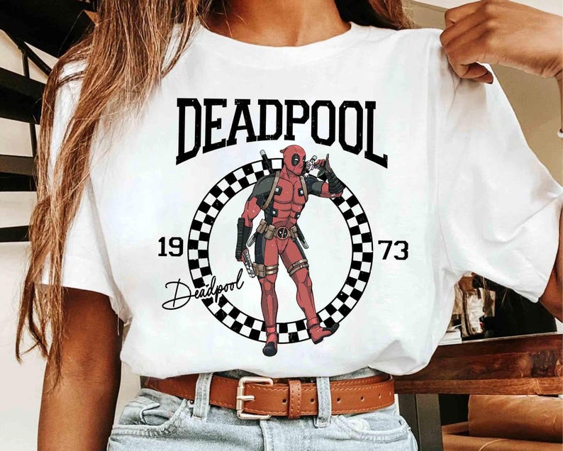 Puede incluir: Camiseta blanca con un dise&ntilde;o de Deadpool. El dise&ntilde;o incluye la palabra "DEADPOOL" sobre un c&iacute;rculo a cuadros con la ilustraci&oacute;n del personaje, los n&uacute;meros "1973" y la firma de Deadpool.