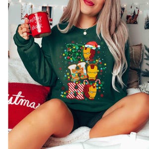 Puede incluir: Una sudadera de manga larga de color verde oscuro con un estampado gr&aacute;fico del texto HO HO HO con m&aacute;scaras de Iron Man integradas en las letras. El dise&ntilde;o incluye luces de Navidad, bastones de caramelo y un gorro de Pap&aacute; Noel. Se sostiene una taza roja.