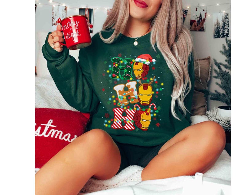 Puede incluir: Una sudadera de manga larga de color verde oscuro con un estampado gr&aacute;fico del texto HO HO HO con m&aacute;scaras de Iron Man integradas en las letras. El dise&ntilde;o incluye luces de Navidad, bastones de caramelo y un gorro de Pap&aacute; Noel. Se sostiene una taza roja.