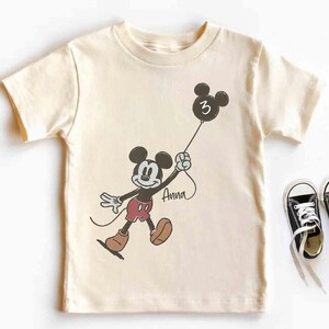 Puede incluir: Camiseta color crema con un dibujo de Mickey Mouse sosteniendo un globo con el n&uacute;mero 3. El nombre "Anna" est&aacute; escrito debajo. Zapatillas negras con cordones blancos junto a la camiseta. Un dise&ntilde;o divertido con tem&aacute;tica de cumplea&ntilde;os.