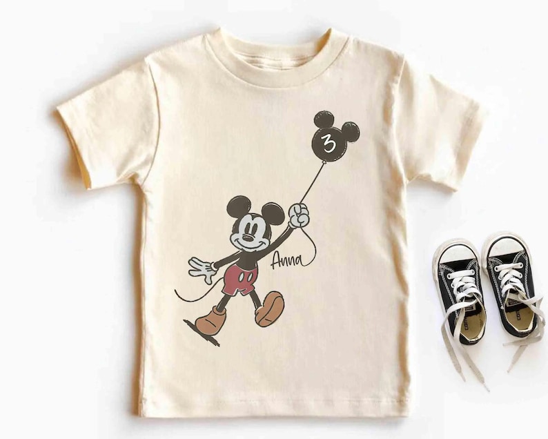 Puede incluir: Camiseta color crema con un dibujo de Mickey Mouse sosteniendo un globo con el n&uacute;mero 3. El nombre "Anna" est&aacute; escrito debajo. Zapatillas negras con cordones blancos junto a la camiseta. Un dise&ntilde;o divertido con tem&aacute;tica de cumplea&ntilde;os.