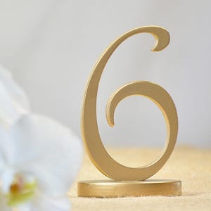 May include: A gold-colored table number six featuring a stylized, curved numeral design mounted on a circular base, positioned next to white orchid flowers.