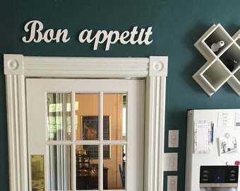 May include: A white door with a glass pane and a sign above it that reads "Bon appétit". The door is in a room with teal walls and a wooden table and chair.