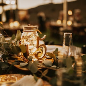 May include: A glass mason jar filled with warm white fairy lights, positioned next to a wooden table number five. The centerpiece is surrounded by green eucalyptus leaves and placed on a round wooden base, creating a soft, glowing display.