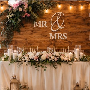 May include: A rustic wooden backdrop featuring the white text MR & MRS, adorned with a large floral arrangement and string lights. A table draped in white fabric sits in front, decorated with a long floral centerpiece, candles, and lanterns.
