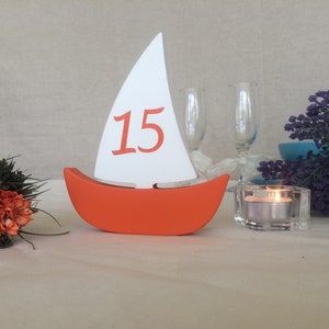 May include: Orange and white wooden sailboat table number with the number 15 on the sail. The sailboat is sitting on a white tablecloth.