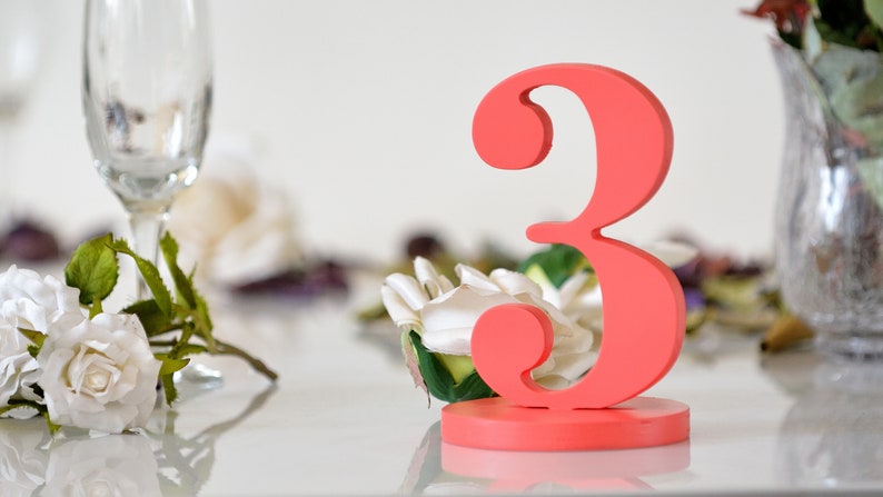 May include: A coral-colored wooden number three table sign featuring a smooth, matte finish and a sturdy circular base. The numeral is upright and stands independently, showcasing a bold, curved font style with a vibrant, solid pigment.