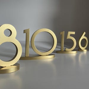 May include: A set of gold-colored table numbers featuring the digits 8, 10, 15, and 6. Each number is constructed with a smooth, matte finish and stands upright on a matching oval-shaped base, providing a stable display for event table identification.