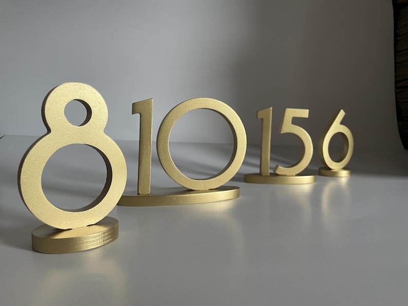 May include: A set of gold-colored table numbers featuring the digits 8, 10, 15, and 6. Each number is constructed with a smooth, matte finish and stands upright on a matching oval-shaped base, providing a stable display for event table identification.