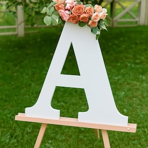 May include: A large white wooden letter "A" on a wooden easel, adorned with a bouquet of orange and pink roses and greenery. The sign is set on a green lawn, suggesting an event or celebration.