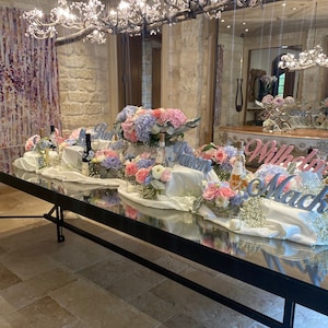 May include: A long, rectangular table with a mirrored surface is set for a wedding reception. The table is decorated with white linens, flowers, and candles. The flowers are in shades of pink, purple, and white. The table is set with white plates, silverware, and glasses. The words "Will You Marry Me" are spelled out in large letters on the table.