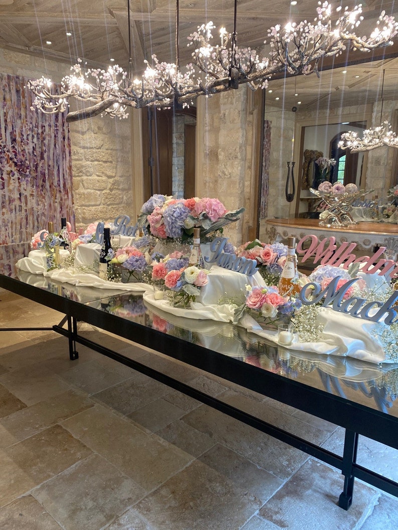 May include: A long, rectangular table with a mirrored surface is set for a wedding reception. The table is decorated with white linens, flowers, and candles. The flowers are in shades of pink, purple, and white. The table is set with white plates, silverware, and glasses. The words "Will You Marry Me" are spelled out in large letters on the table.
