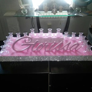 May include: A pink tiered cake stand with a sparkly silver trim. The stand is decorated with the name "Gl&oacute;rissa" in pink letters. There are clear glass candle holders on each tier.