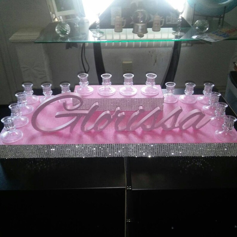 May include: A pink tiered cake stand with a sparkly silver trim. The stand is decorated with the name "Gl&oacute;rissa" in pink letters. There are clear glass candle holders on each tier.