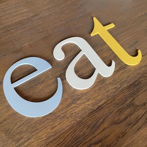 May include: The word "eat" spelled out in three dimensional letters. The letter "e" is light blue, the letter "a" is white, and the letter "t" is yellow.