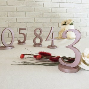 May include: A collection of freestanding table numbers painted in a metallic rose gold finish. The numbers 10, 5, 8, 4, and 3 are displayed, each featuring a smooth, rounded font and a circular base for stability on a flat surface.