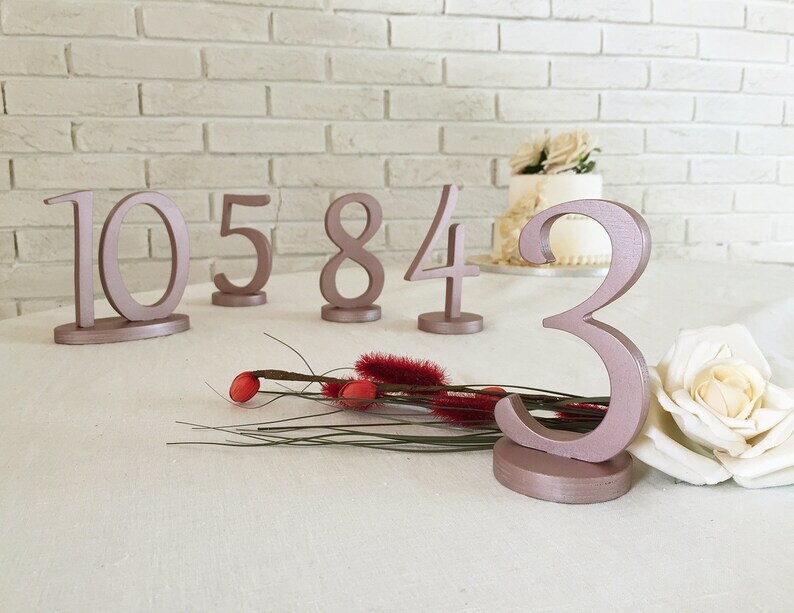 May include: A collection of freestanding table numbers painted in a metallic rose gold finish. The numbers 10, 5, 8, 4, and 3 are displayed, each featuring a smooth, rounded font and a circular base for stability on a flat surface.