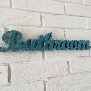 May include: Teal blue sign with the word "Bathroom" in a cursive font, mounted on a white brick wall.