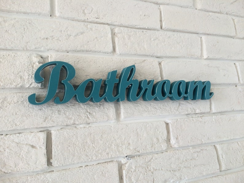 May include: Teal blue sign with the word "Bathroom" in a cursive font, mounted on a white brick wall.