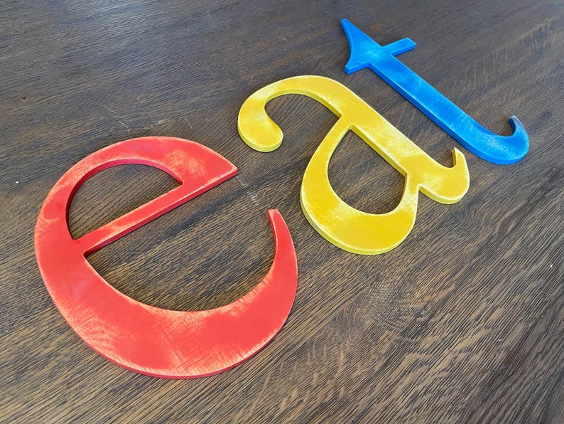 May include: Three wooden letters spelling out the word "Eat" in red, yellow, and blue paint. The letters are arranged horizontally on a wooden surface.