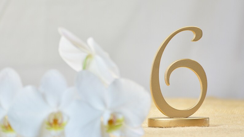 May include: A gold-colored table number sign in the shape of the numeral six, featuring a smooth finish and a circular base, positioned next to white orchid flowers with yellow centers, creating a clear visual focus on the metallic number.