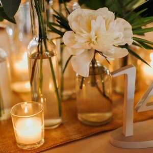 May include: A white table number 14 sign with an oval base, positioned next to clear glass bottles containing white flowers and green foliage, accompanied by a small glass votive candle holder with a lit candle on a textured tan runner.