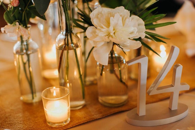 May include: A white table number 14 sign with an oval base, positioned next to clear glass bottles containing white flowers and green foliage, accompanied by a small glass votive candle holder with a lit candle on a textured tan runner.
